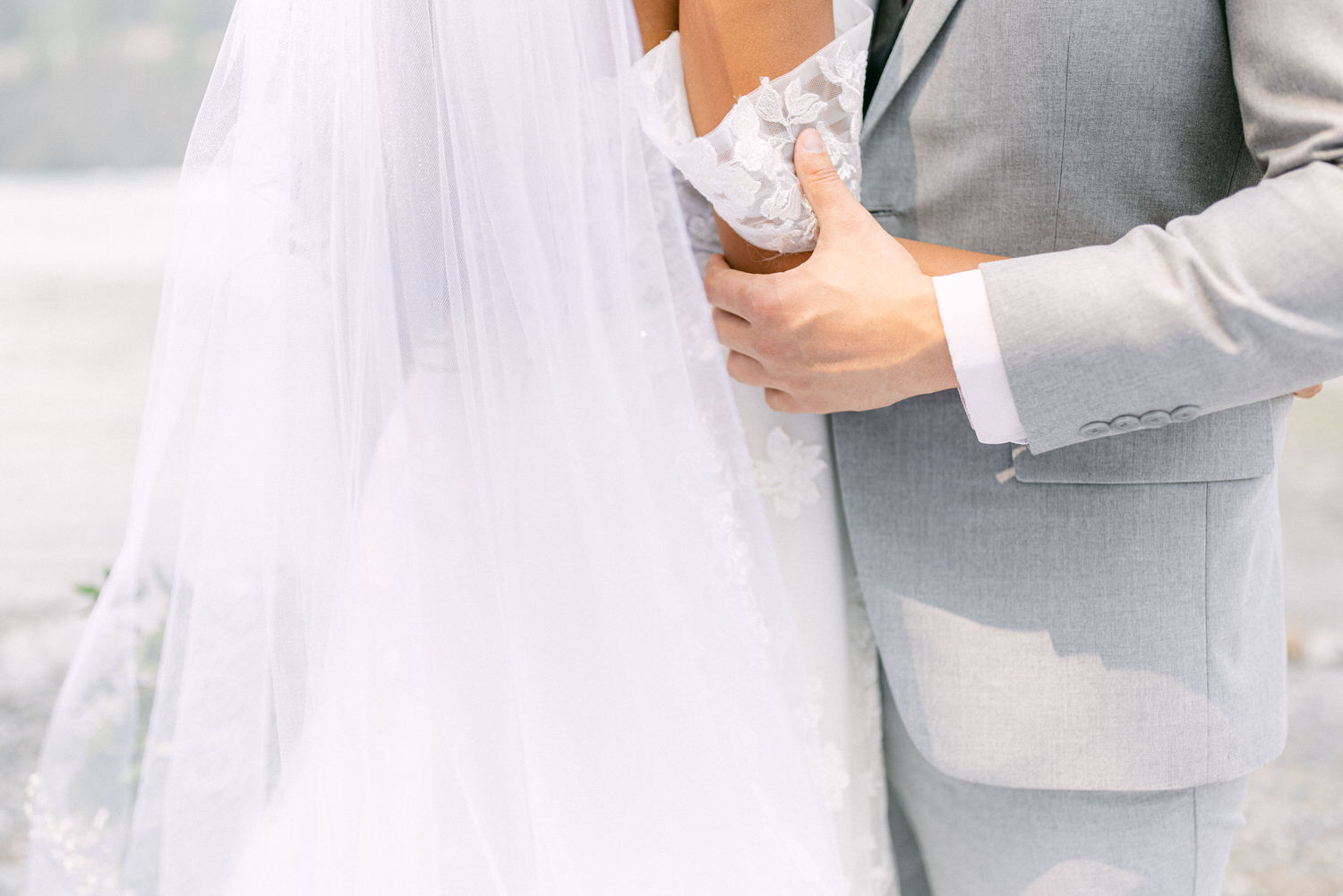 A close-up of a couple in wedding attire, showcasing the groom's elegant gray suit and the bride's delicate lace sleeve and flowing veil, amidst a scenic backdrop.