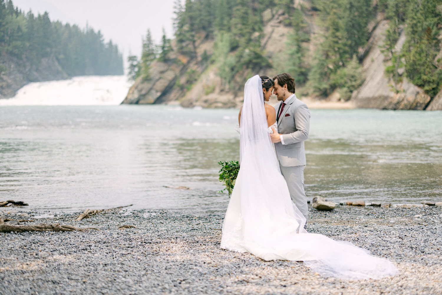 Couple embracing at the edge of a serene lake with a waterfall in the background, surrounded by trees.