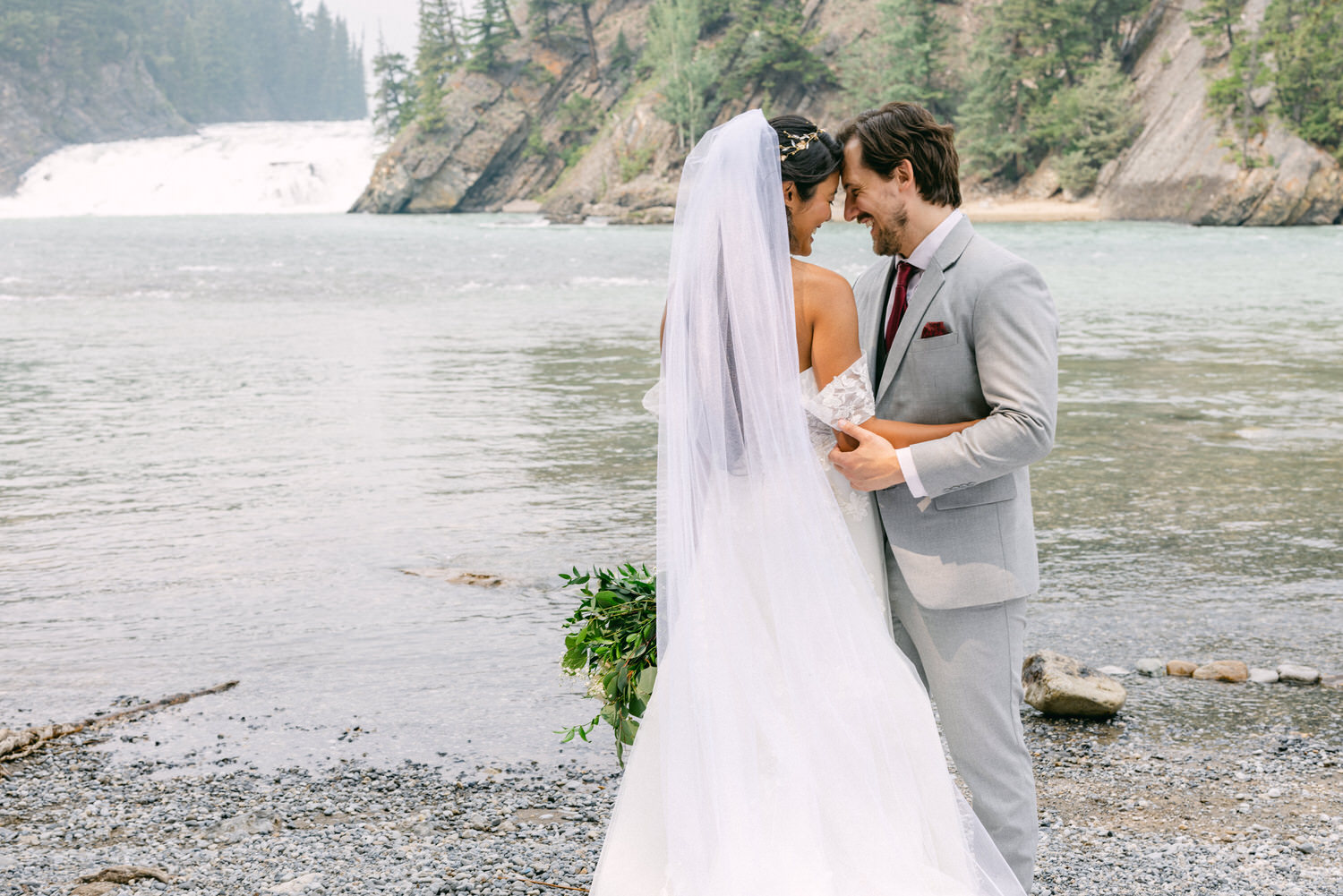 A couple sharing a romantic moment by a serene river, with a waterfall in the background, as the bride holds her bouquet and they embrace intimately.