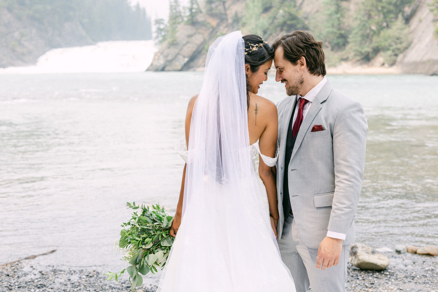 A bride and groom share a tender moment by a serene river, surrounded by lush greenery and a distant waterfall. The bride holds a bouquet of fresh greenery, and both wear elegant attire.