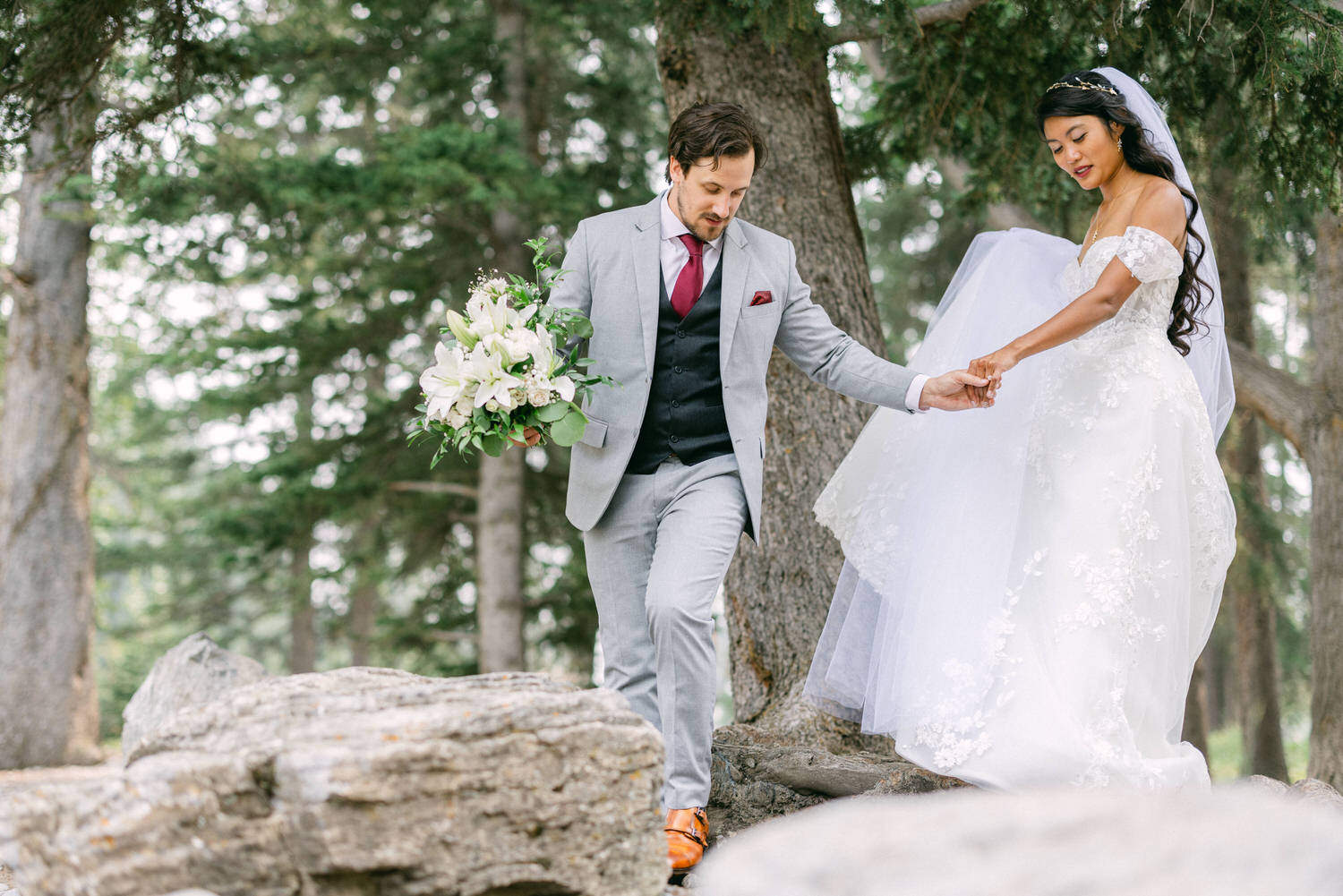 A groom in a light gray suit assists his bride, who is wearing a white wedding gown, as they navigate through rocky terrain in a forest setting. The bride holds a bouquet of white flowers, while trees surround them.