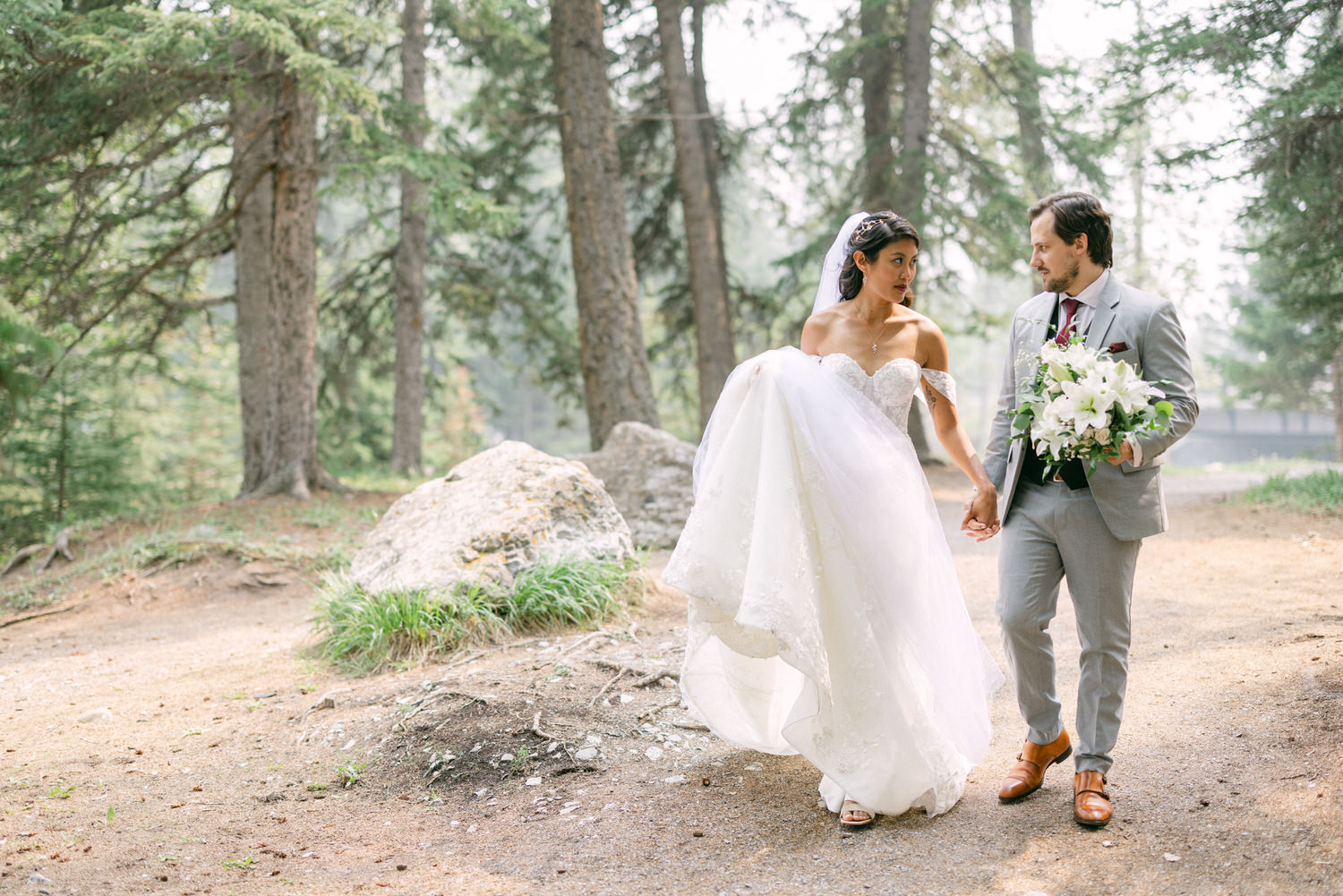 Bride and groom walking hand in hand through a wooded area, with the bride in a flowing wedding dress and the groom holding a bouquet.