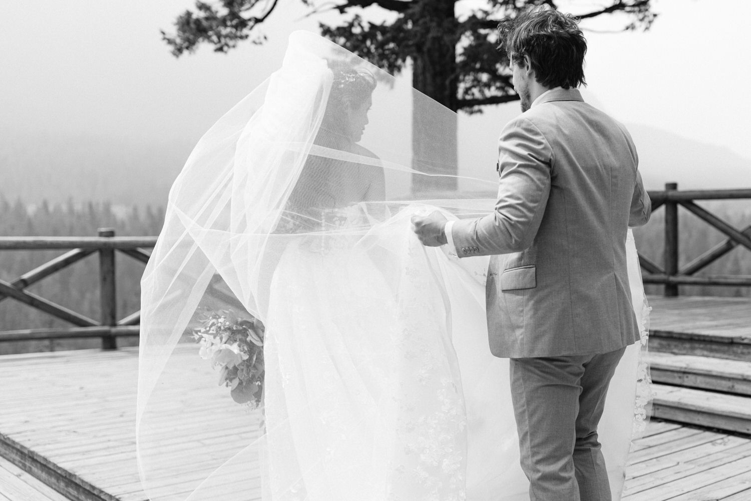 A groom gently lifts the veil of a bride on a misty outdoor deck, capturing a tender moment before their wedding.