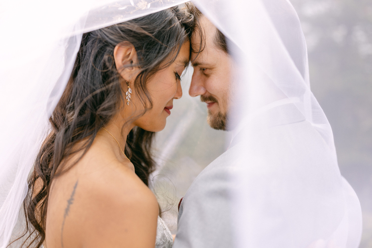 A couple sharing a tender moment under a wedding veil, gazing into each other's eyes, surrounded by a soft, dreamy atmosphere.