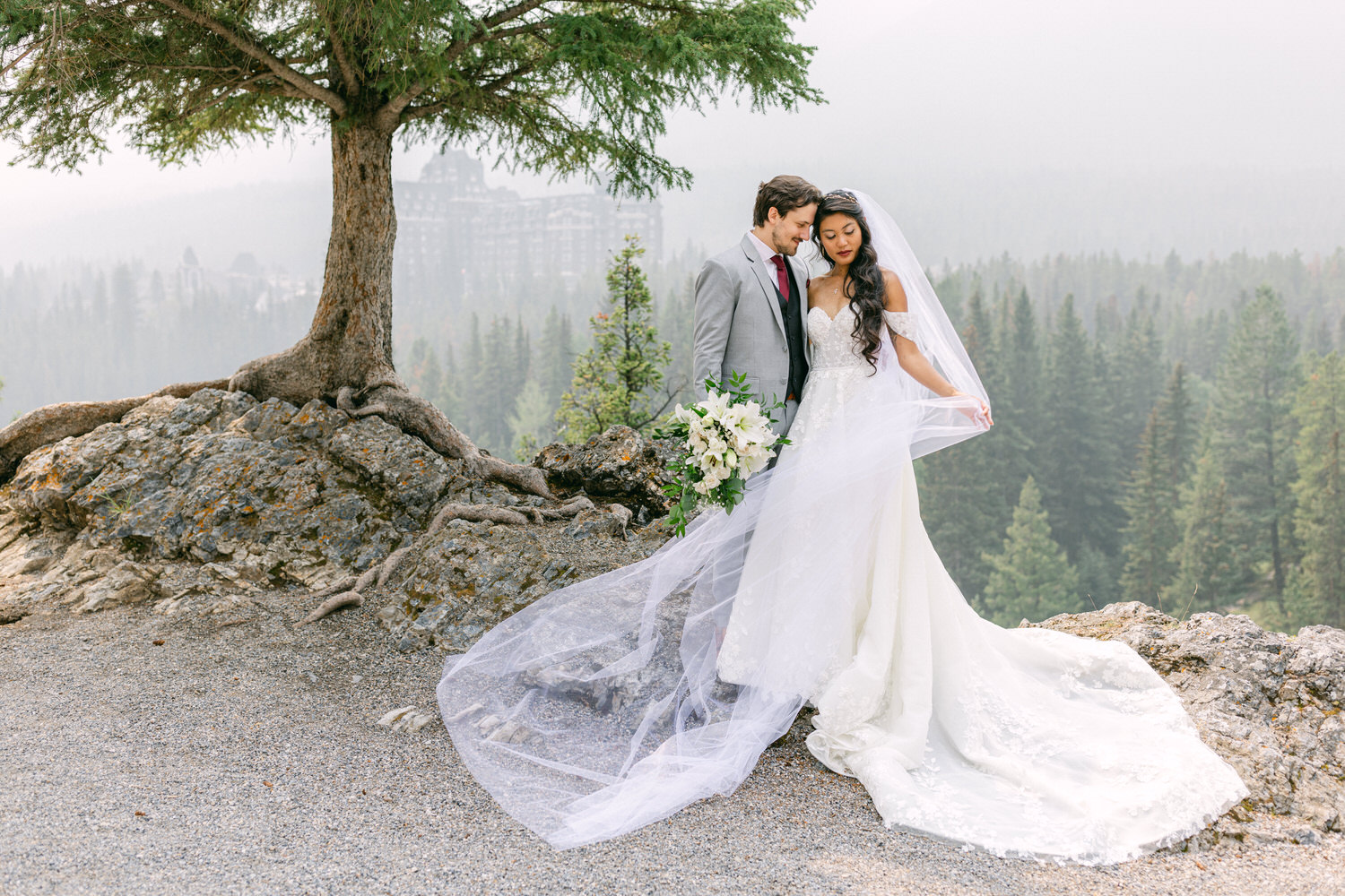 A bride and groom surrounded by a misty forest, embracing with a scenic backdrop of trees and a distant lodge.