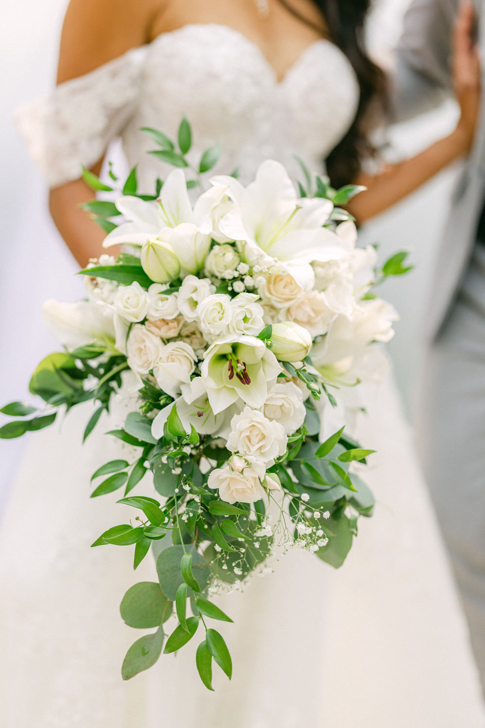 A close-up of a beautiful bridal bouquet featuring white lilies, roses, and greenery, held by a bride in a lace dress.