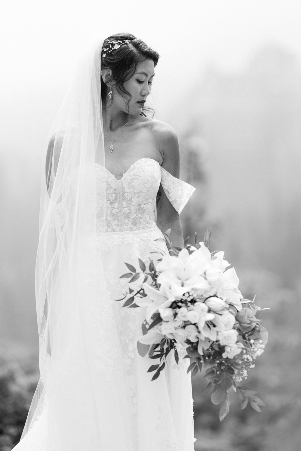 A poised bride wearing a delicate lace gown and veil, holding a bouquet of white flowers, set against a soft, foggy backdrop.