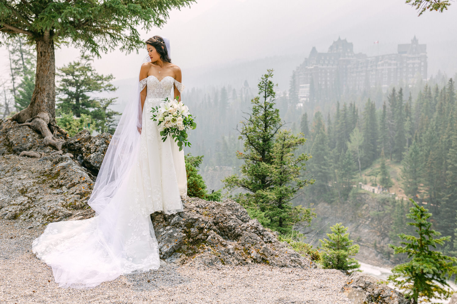 A bride in an elegant gown with a long veil stands on a rocky outcrop, holding a bouquet of white flowers against a misty forest backdrop.