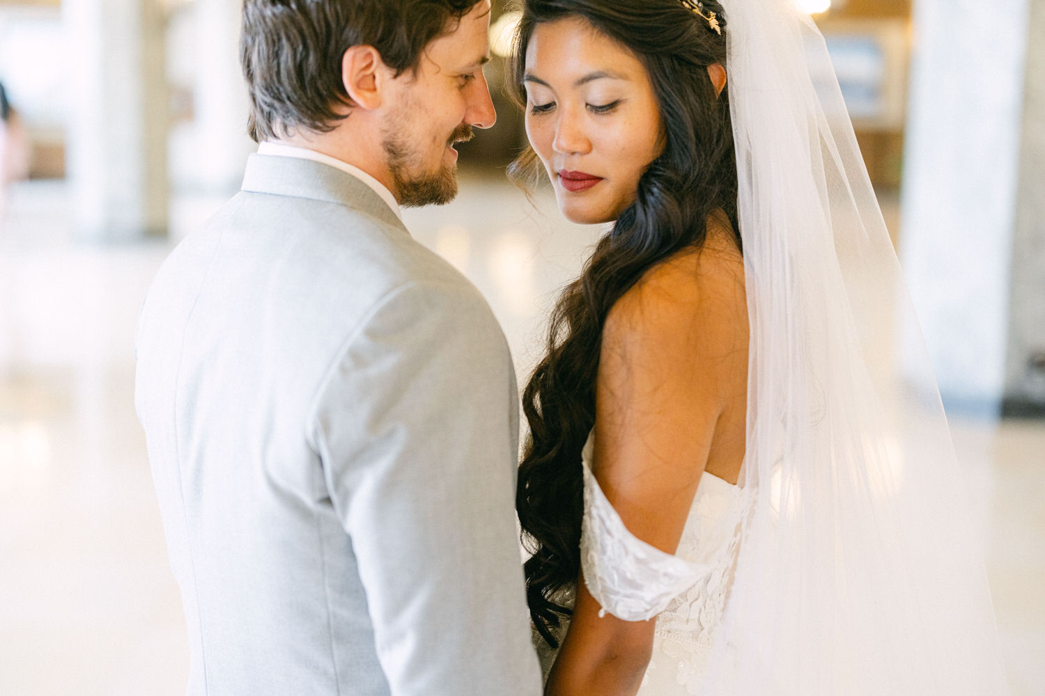 A close-up of a bride and groom, gazing lovingly at each other, with the bride wearing a white gown and veil, and the groom in a light gray suit, set against a softly lit background.