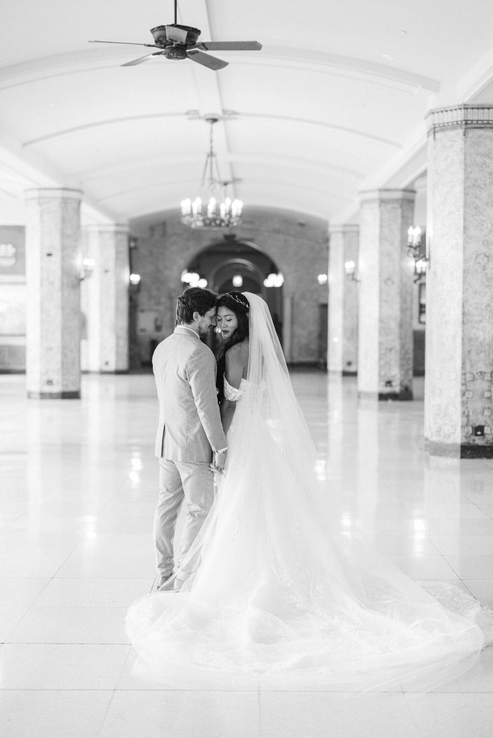 A couple shares an intimate moment in a grand, empty hall, surrounded by elegant architecture and soft natural light.