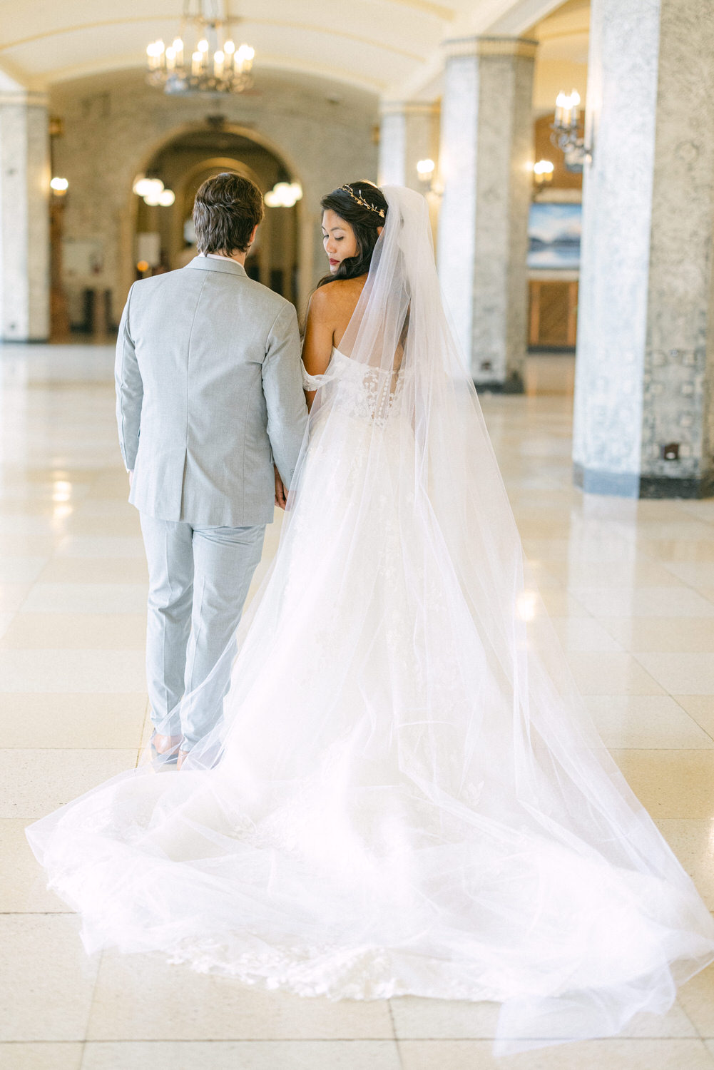 A bride and groom holding hands, walking together down a beautifully lit hallway, dressed in elegant wedding attire. The bride's long veil trails behind her, enhancing the romantic atmosphere.