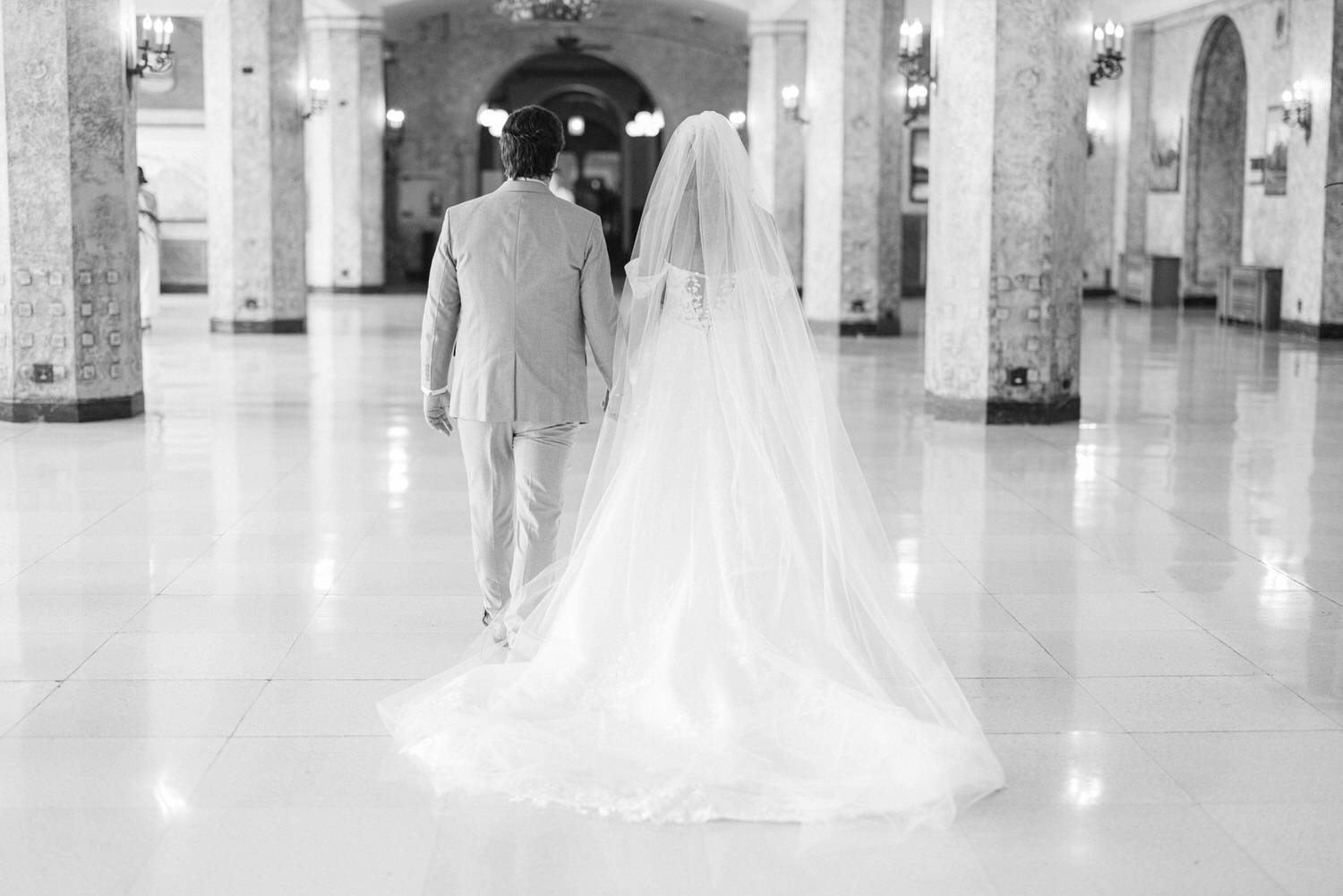 A couple walking hand in hand down a grand, empty hall, the bride in a flowing white gown and veil, and the groom in a light-colored suit.