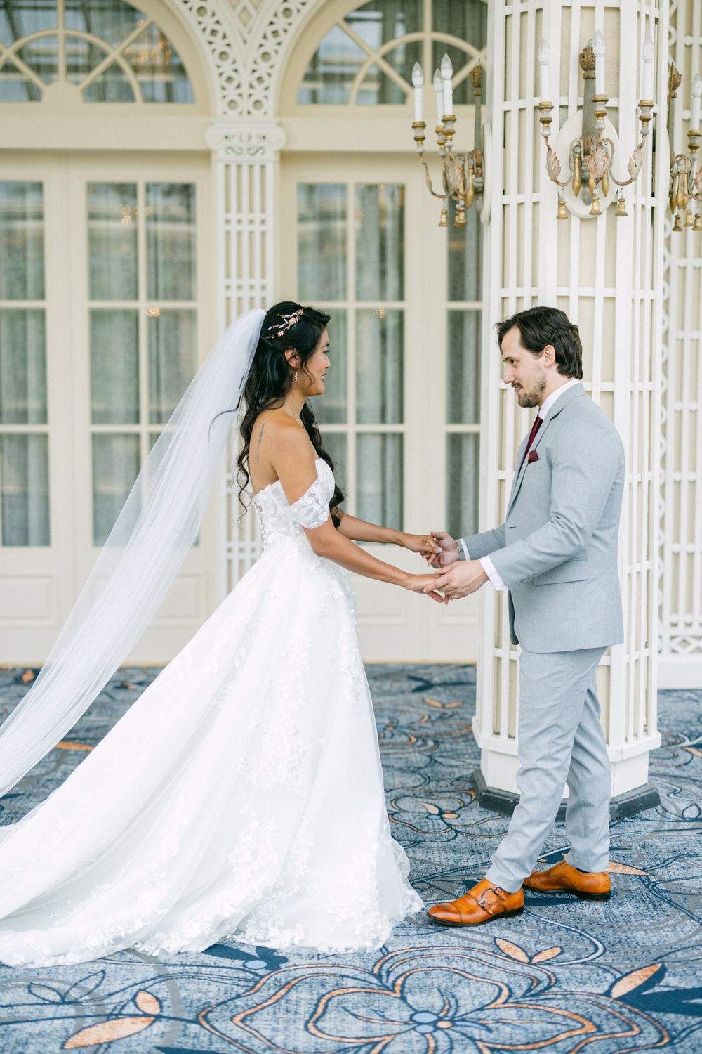 A bride in a beautiful white gown and veil gazes lovingly at her groom in a light gray suit as they hold hands in an elegant indoor setting.