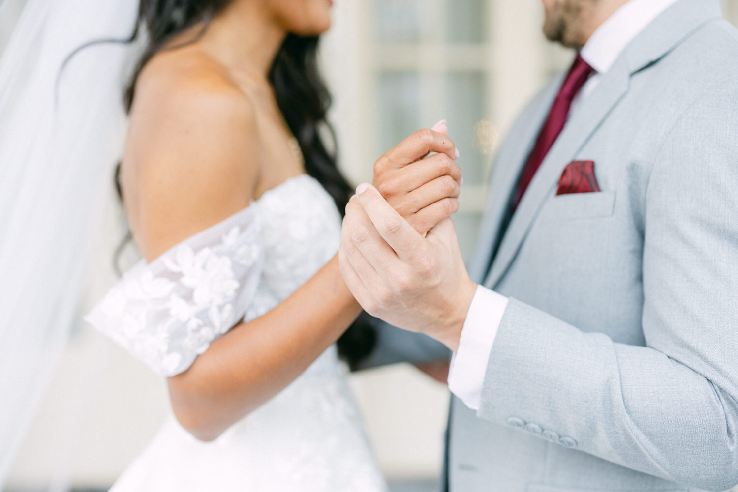 A close-up of a bride and groom holding hands, showcasing their affection and wedding attire, with the bride in a white dress and the groom in a light gray suit.