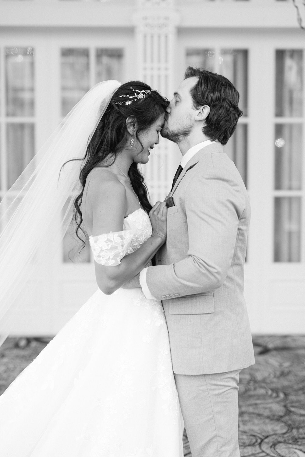 A bride and groom share a tender moment, forehead to forehead, in an elegant setting. The bride is wearing a beautiful white gown with lace detailing, while the groom is in a light gray suit. The ambiance is romantic and intimate, highlighting their joy and love.