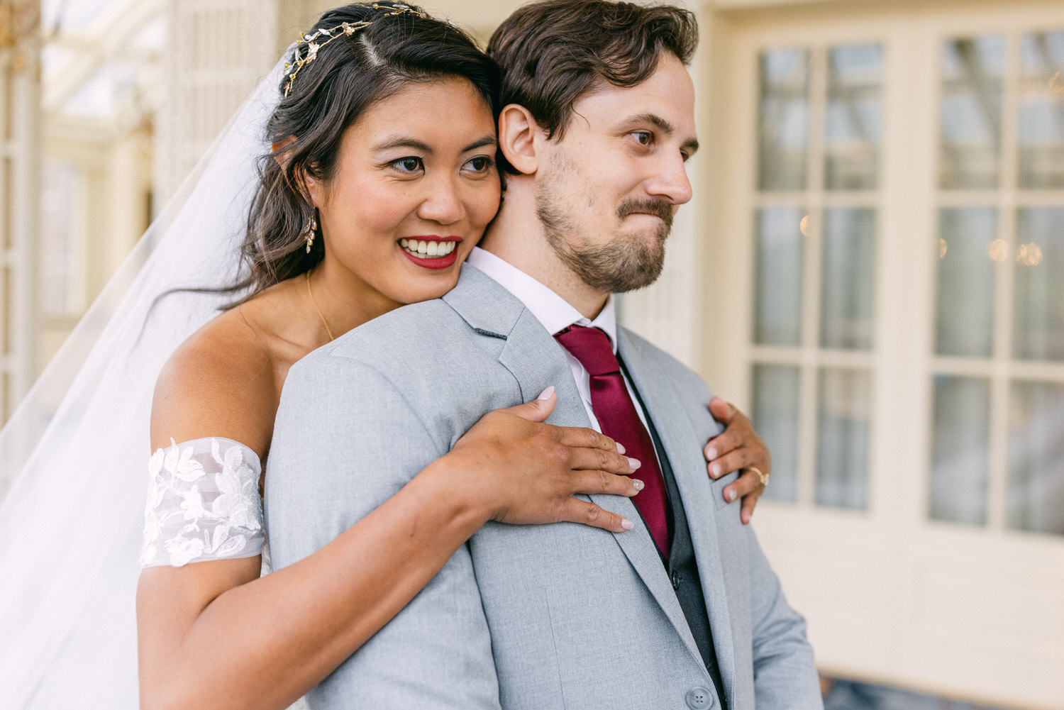 A couple smiles happily in a romantic embrace, showcasing the bride's elegant off-shoulder dress and the groom's sharp suit, set against a beautifully lit backdrop.