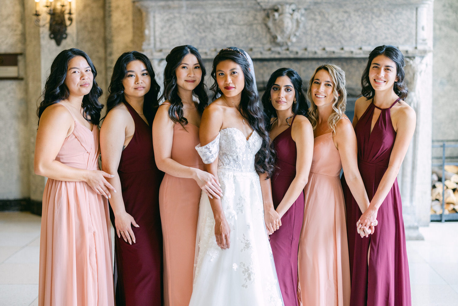 A stunning bridal party poses together, showcasing a bride in a detailed white gown and bridesmaids in soft pink and burgundy dresses against a beautiful ornate background.