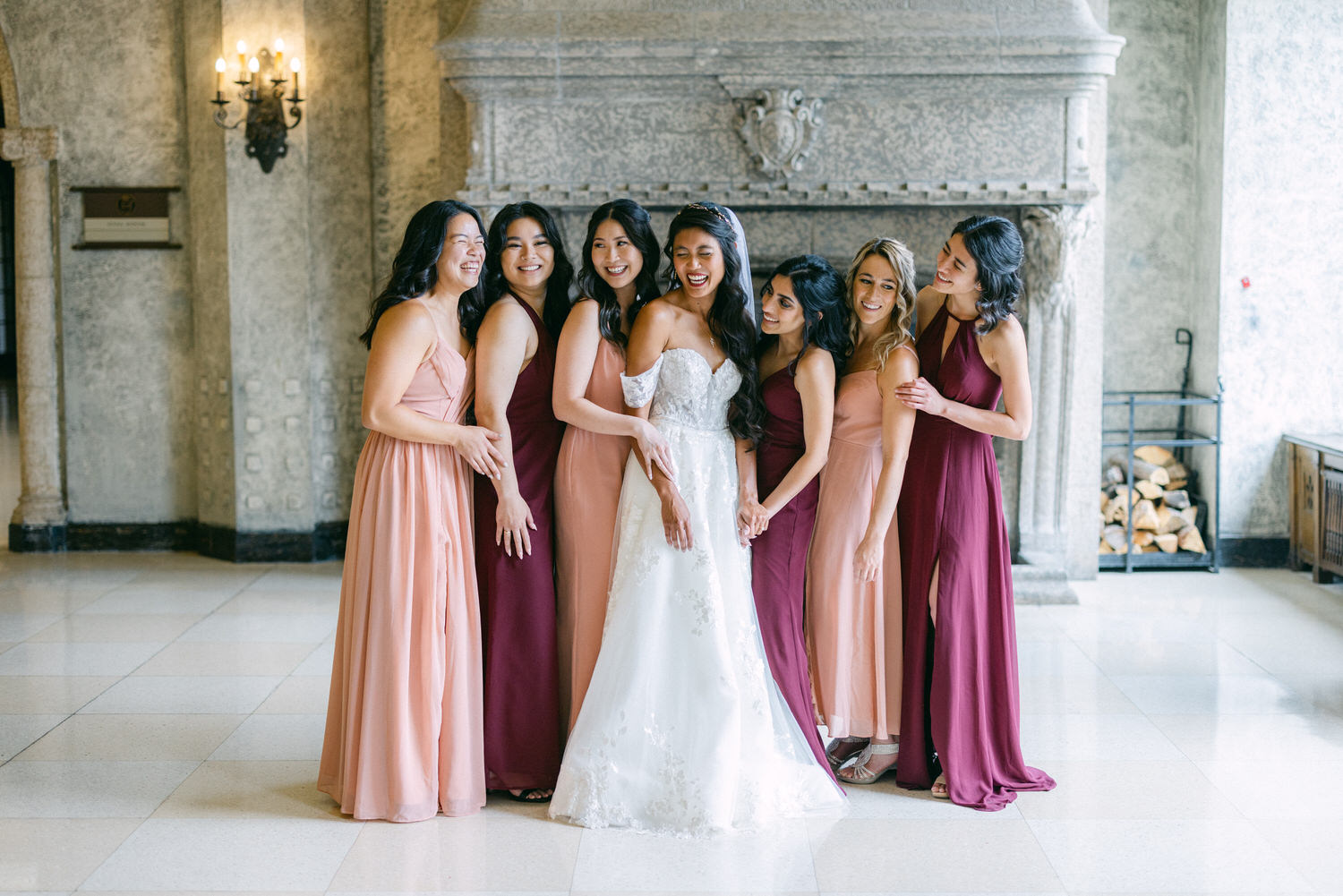 A happy bride surrounded by her bridesmaids, dressed in varying shades of pink and burgundy, in an elegant indoor setting.