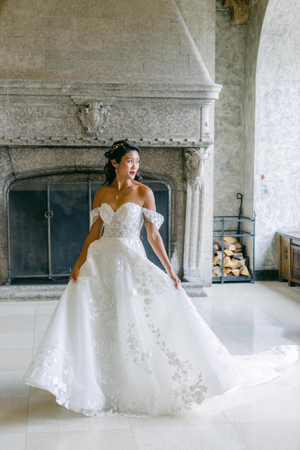 A bride poses gracefully in an elegant off-shoulder wedding gown adorned with floral details, standing in a beautifully styled interior.