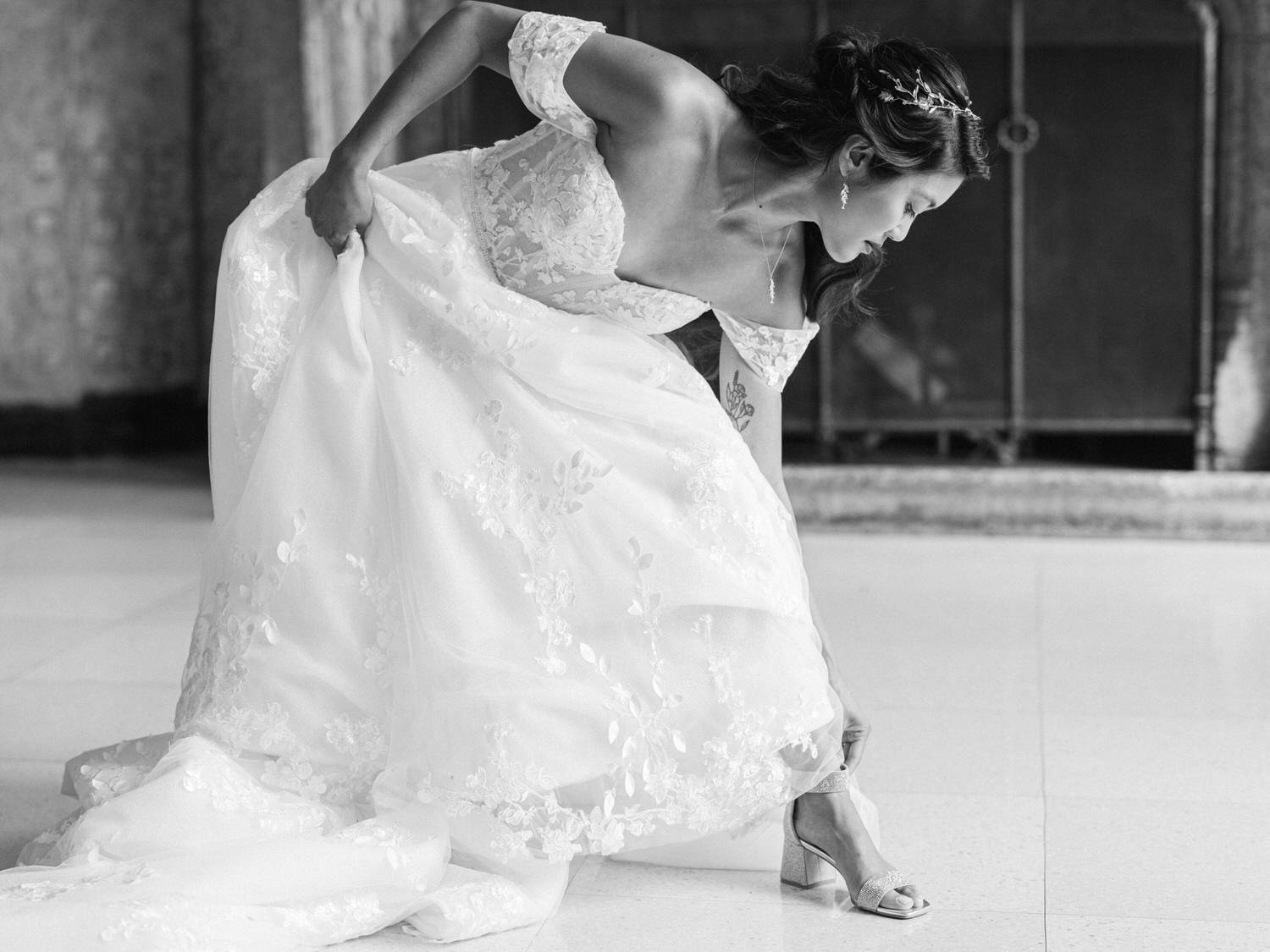 A bride in a beautiful lace gown adjusts her dress while putting on her shoes in an elegant indoor setting.