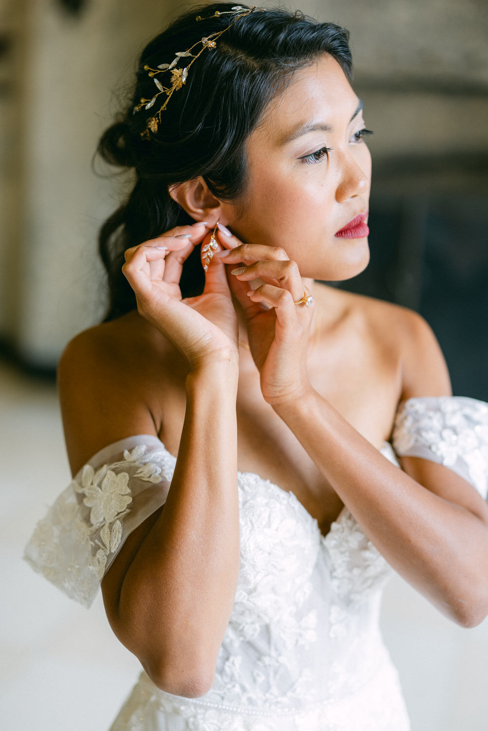 A bride delicately placing her earrings while wearing a stunning off-shoulder wedding gown and a floral hair accessory.