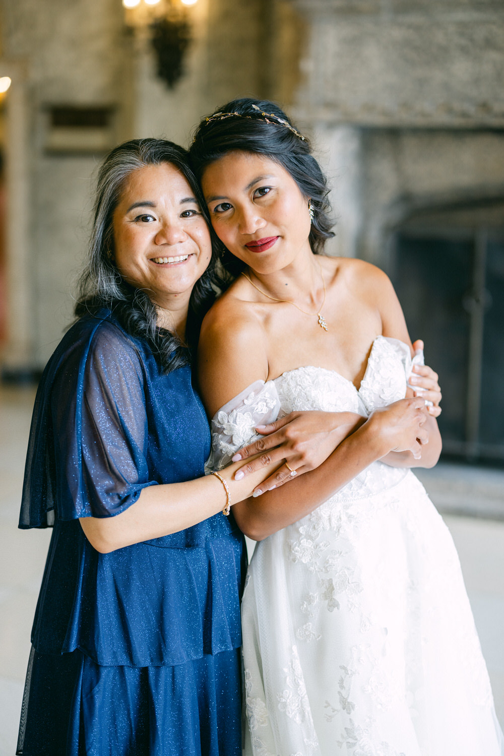 A smiling mother and daughter embrace in a beautiful indoor setting, showcasing the daughter's wedding dress and the mother's elegant attire.