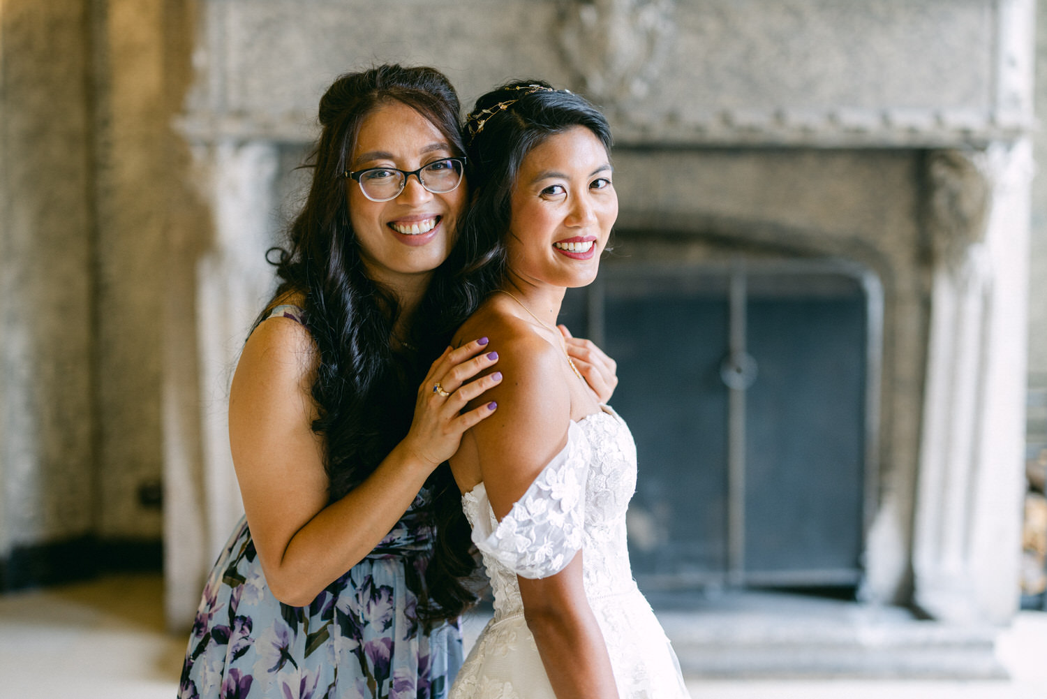 A bride smiles alongside a friend, showcasing their playful and supportive bond, with an elegant background featuring a fireplace.