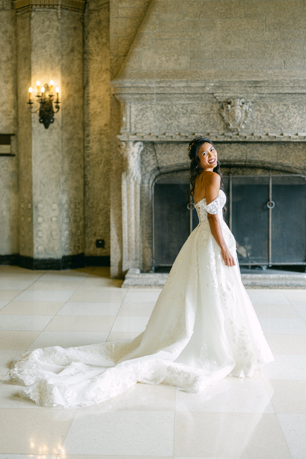 A smiling bride in a flowing white wedding gown twirls gracefully in a lavish historic room, showcasing intricate detailing of her dress against a grand stone fireplace.