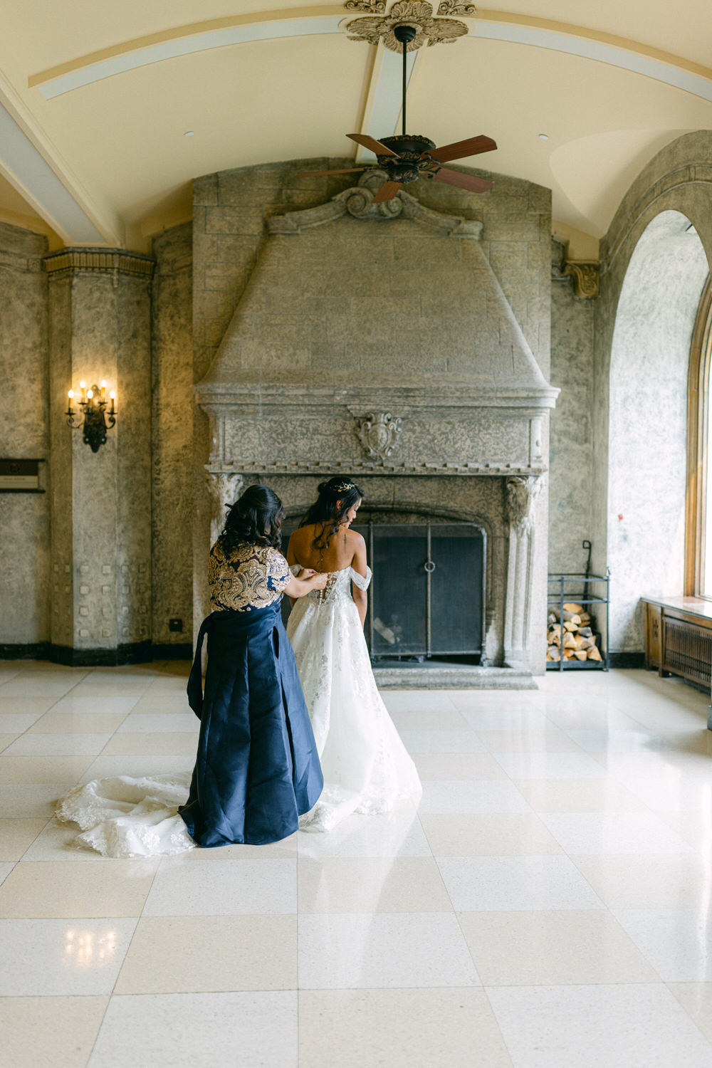 A bride in a wedding gown is assisted by a woman in a richly patterned dress, capturing an intimate moment in an elegant, spacious room with a stone fireplace and large windows.