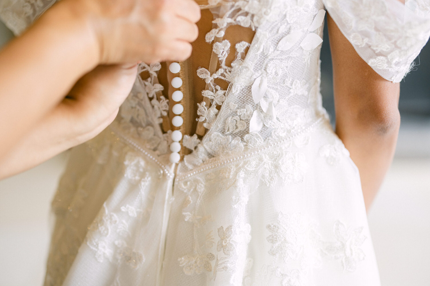 A close-up of a person buttoning the back of a delicate bridal gown adorned with lace and floral embellishments.