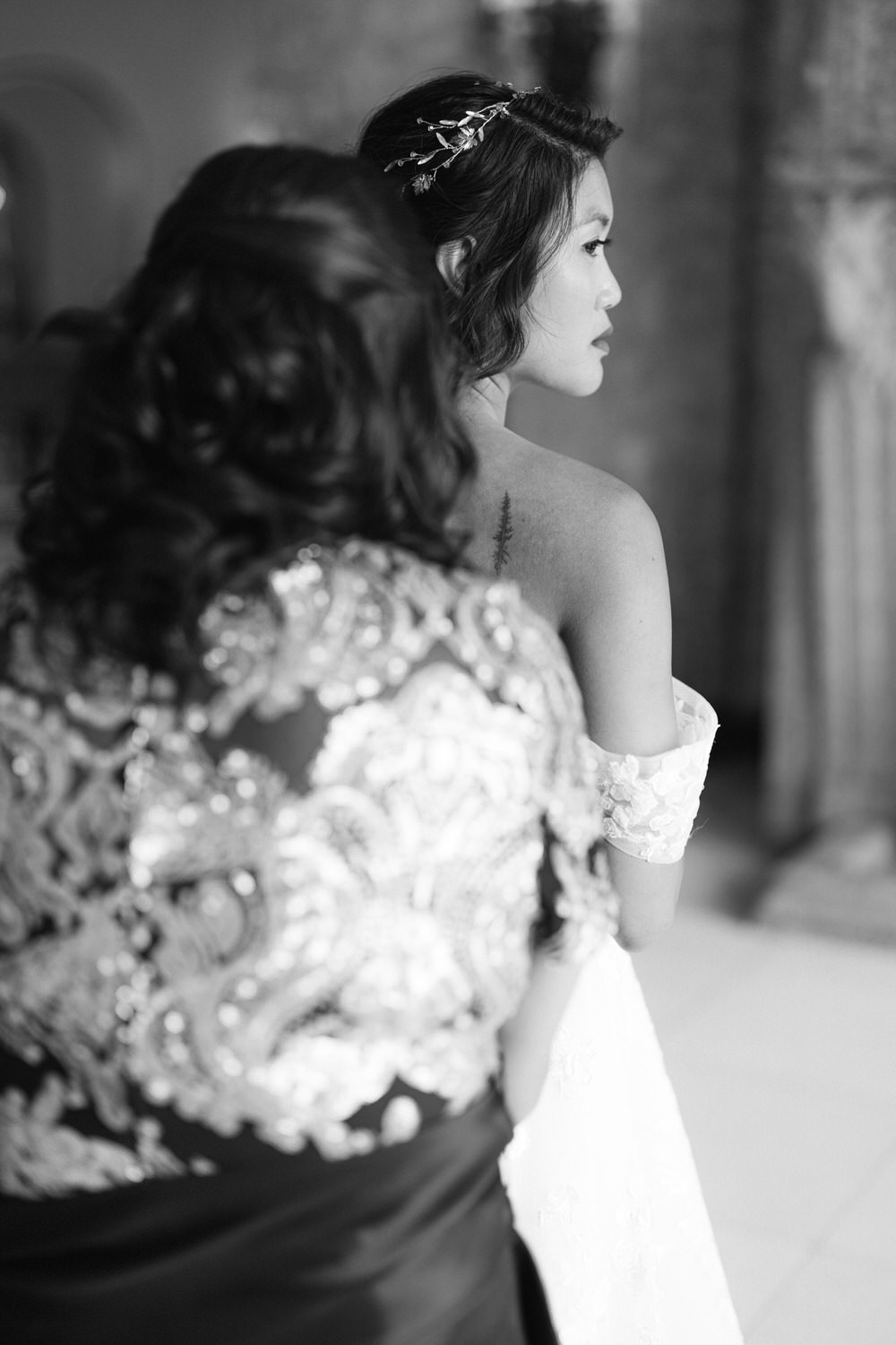 A bride in an elegant gown is assisted by a woman adjusting her attire, captured in a monochrome setting that emphasizes intimacy and detail.