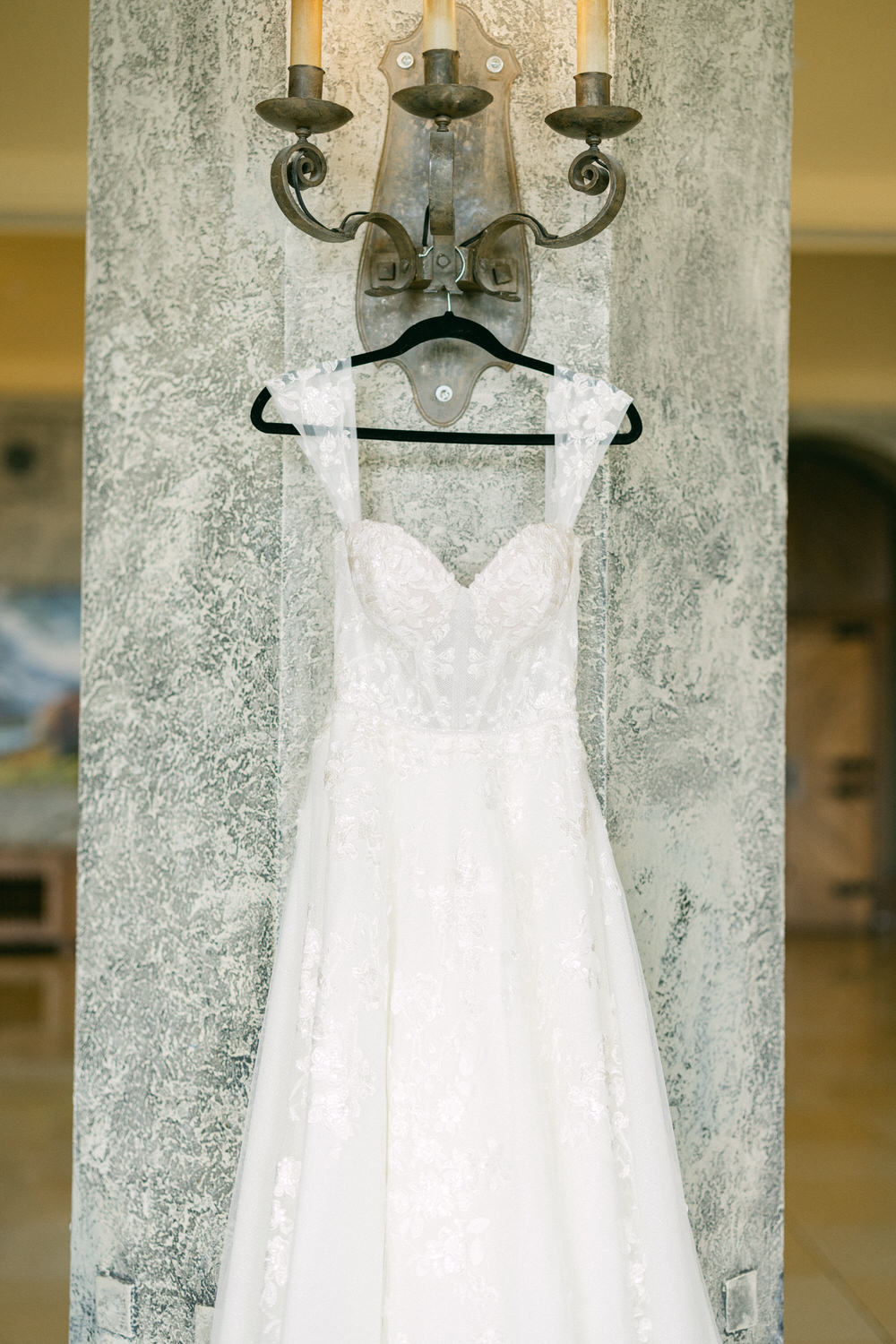 A beautiful wedding dress with a fitted bodice and flowing skirt, hanging on a decorative wall hook, set against a textured wall.