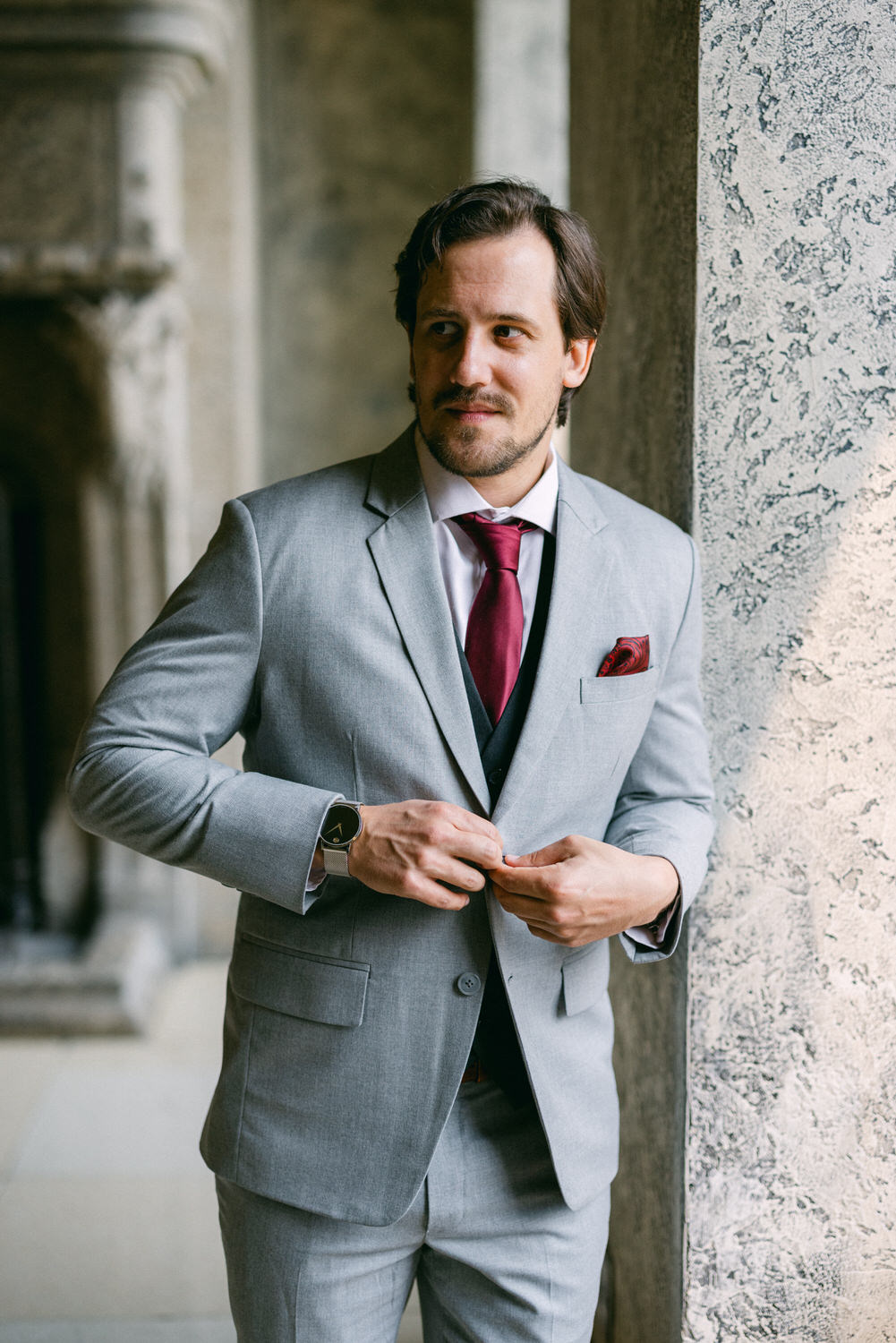 A well-dressed man adjusts his suit jacket, featuring a gray suit and a burgundy tie, standing near a decorative stone wall.