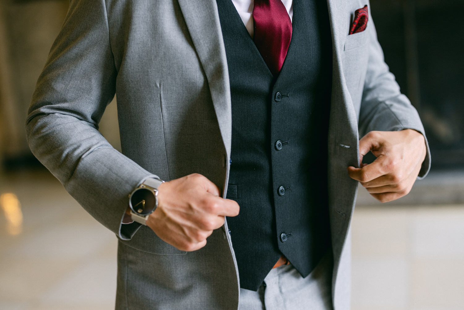 A man in a light gray suit and dark vest adjusts his outfit while showcasing a wristwatch, indicating a polished appearance.