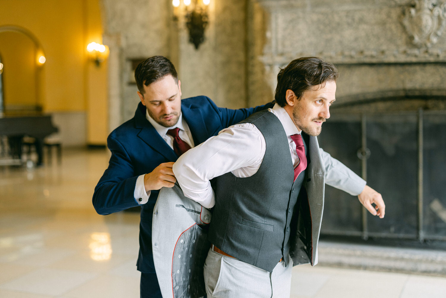 A groom is assisted by a friend as he puts on his suit jacket in an elegant venue, showcasing a moment of camaraderie before the ceremony.