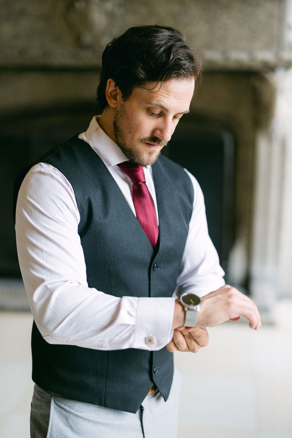 A well-dressed man in a vest and tie adjusting his watch with a focused expression, set against a softly blurred background.