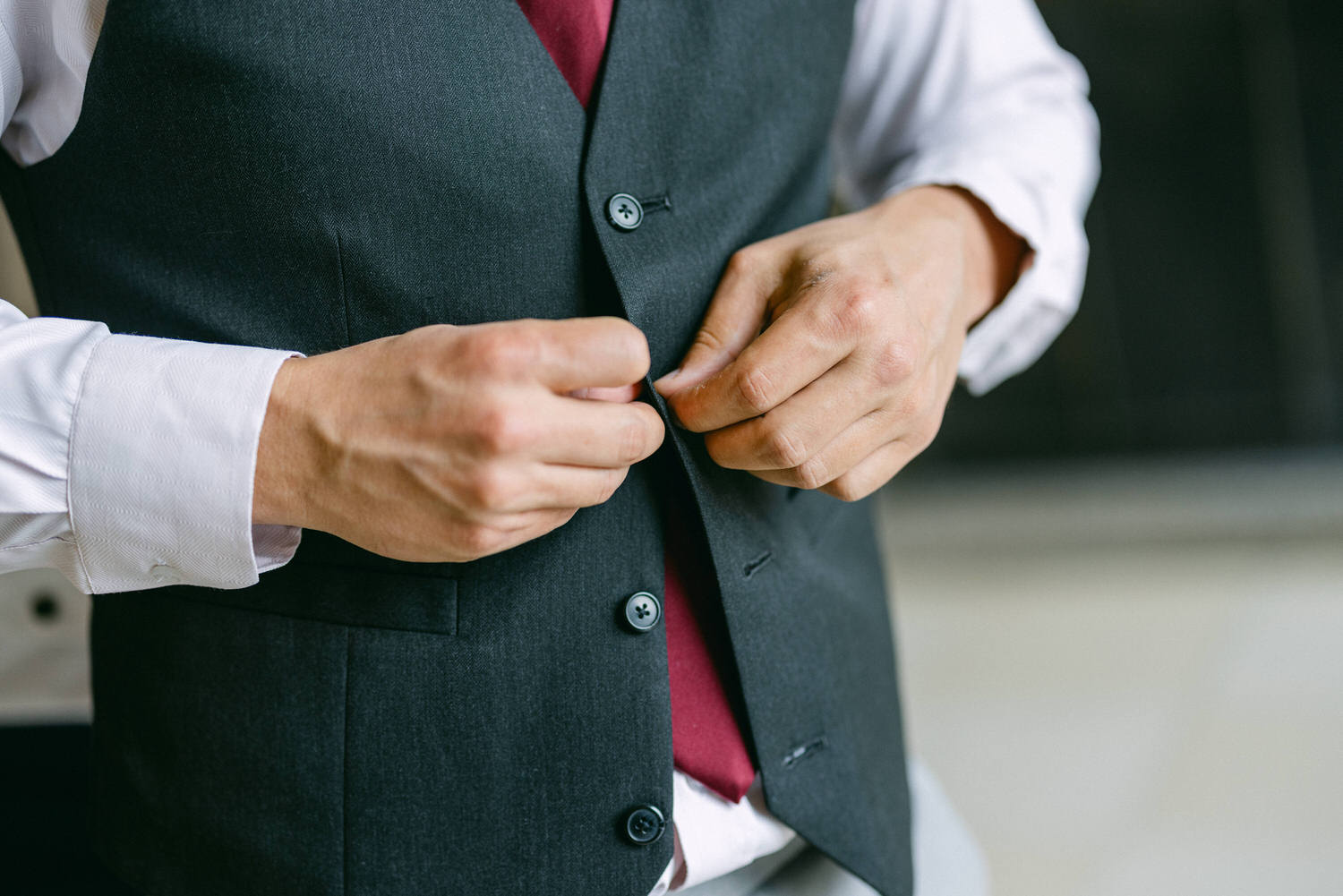 A close-up of a man's hands buttoning a dark vest over a white shirt and maroon tie, capturing the elegance of formal attire.