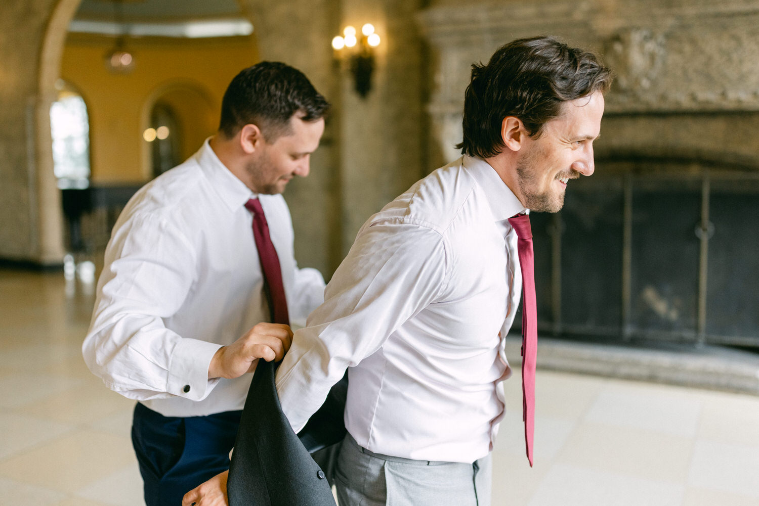 Two men assist each other in putting on suits in an elegant indoor setting.