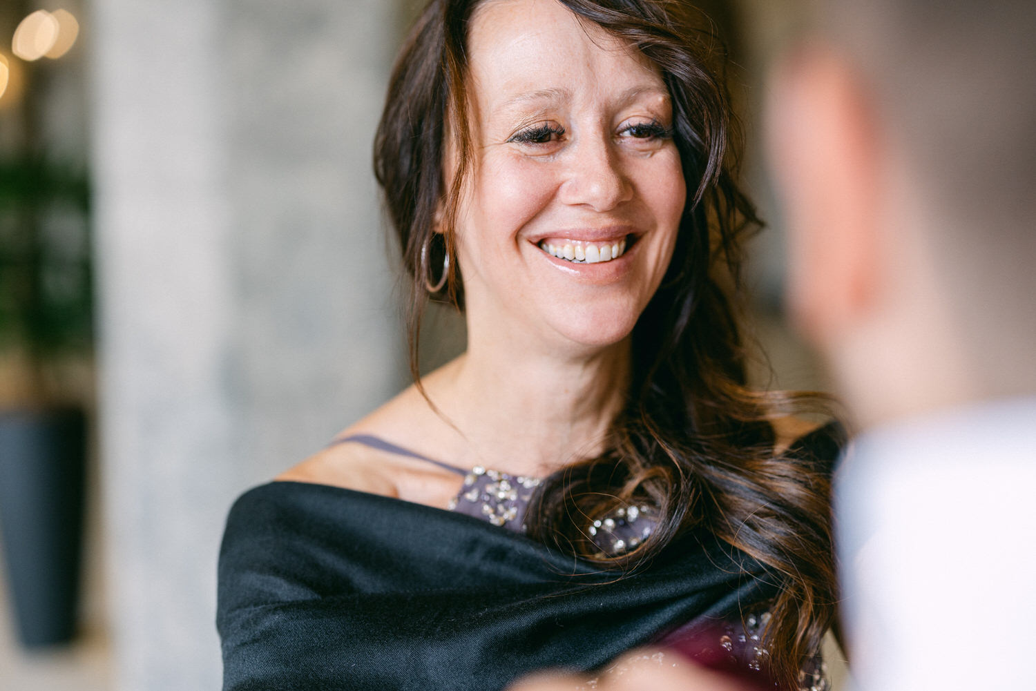 A smiling woman in a black outfit, adorned with sparkling details, enjoying a conversation at a festive event.