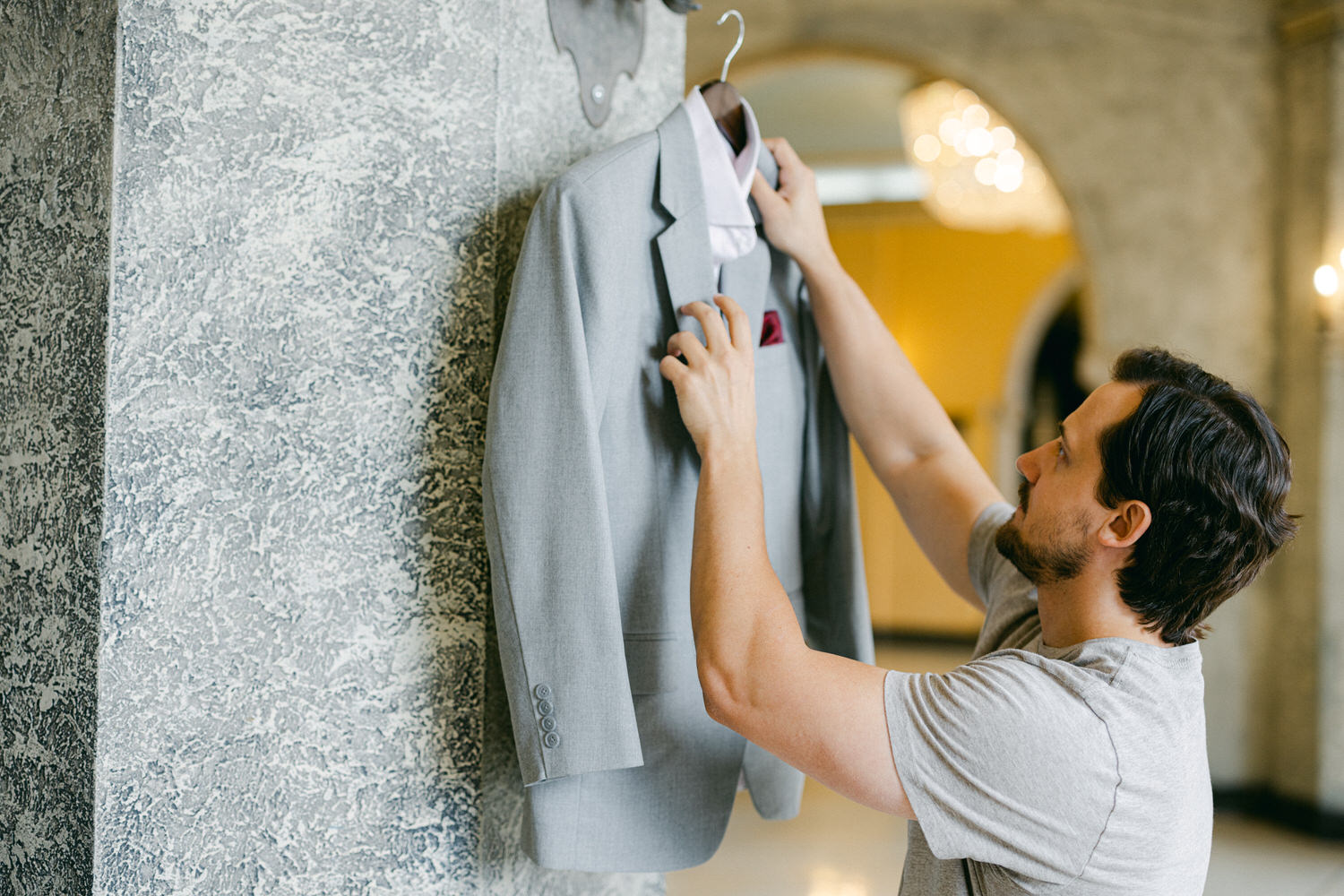 A man adjusts a gray suit jacket hanging on a wall in a stylish indoor setting.