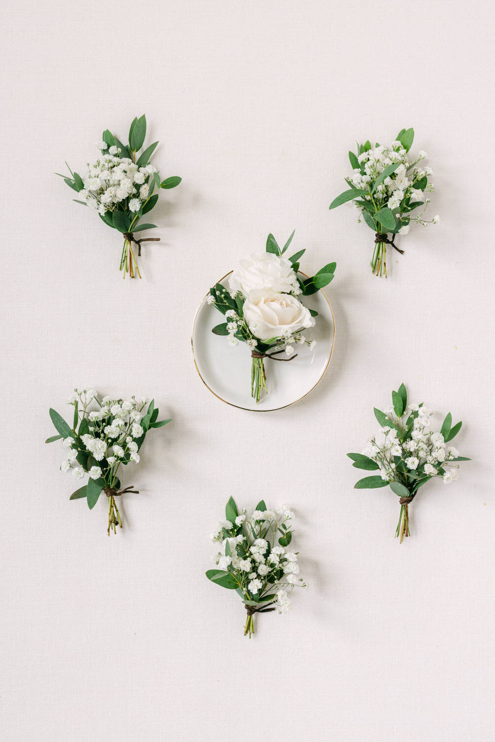 An arrangement of six delicate floral boutonnieres featuring white roses and baby's breath, with one centerpiece on a gold-rimmed plate, set against a soft linen backdrop.