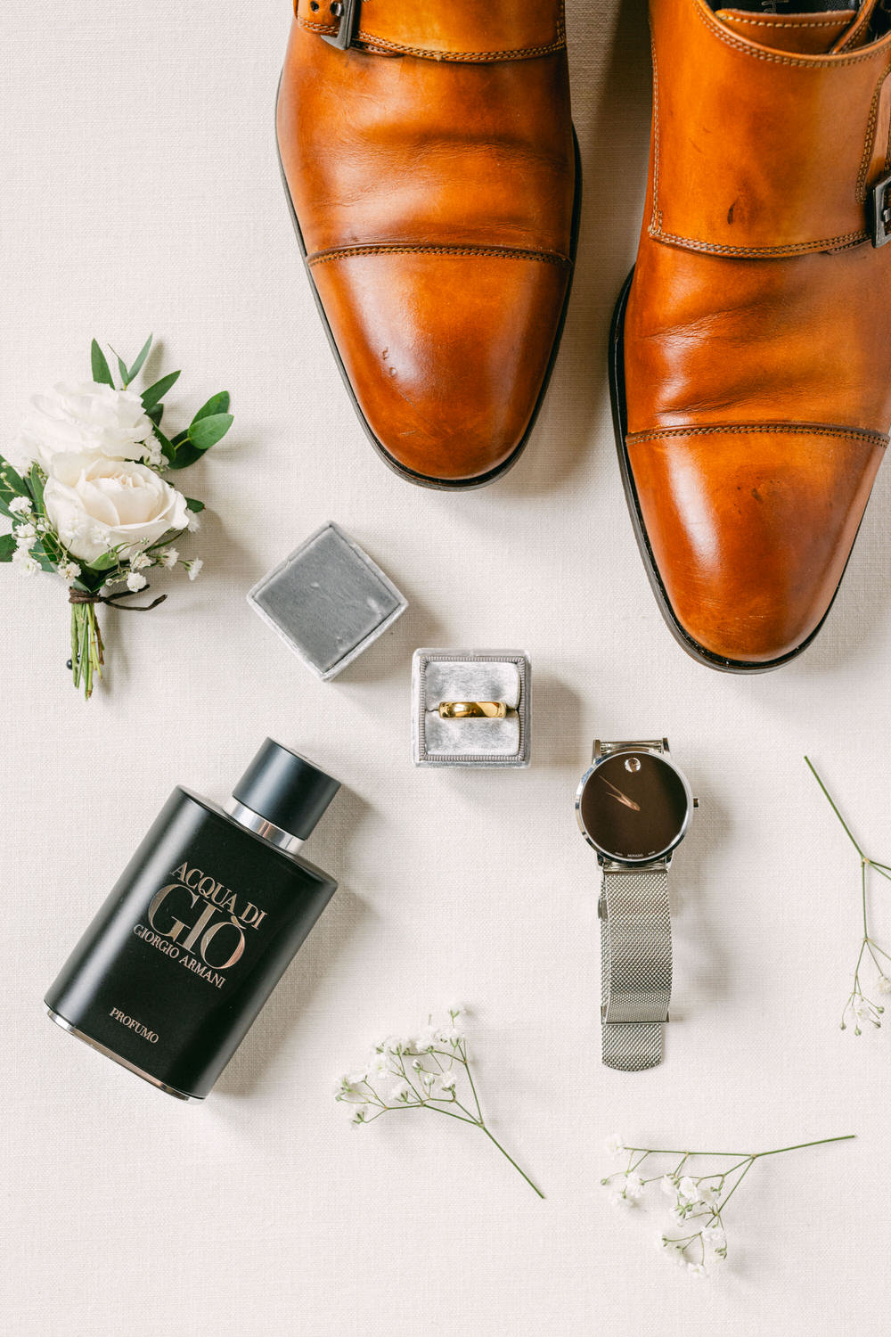 A pair of polished brown shoes, a bottle of Acqua di Gio fragrance, a gold ring in a silver box, a sleek watch, and a boutonniere with white flowers arranged on a neutral fabric background.