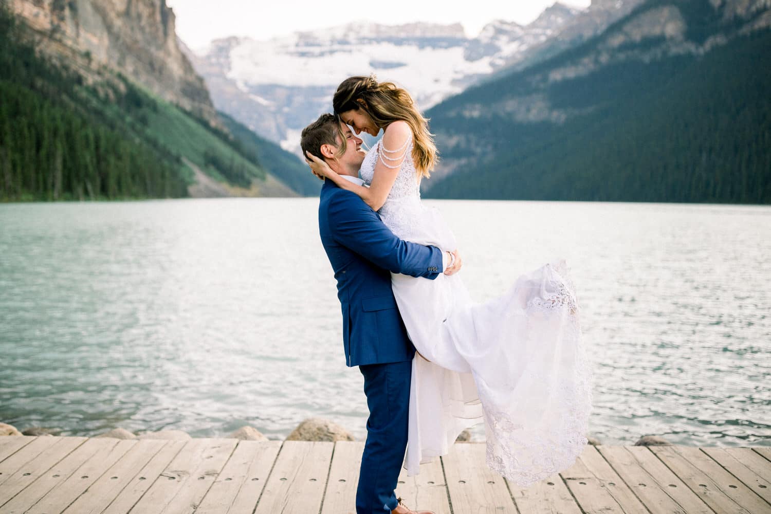 A groom in a blue suit lifts his bride in a white dress, both smiling, with a scenic lake and mountains in the background.