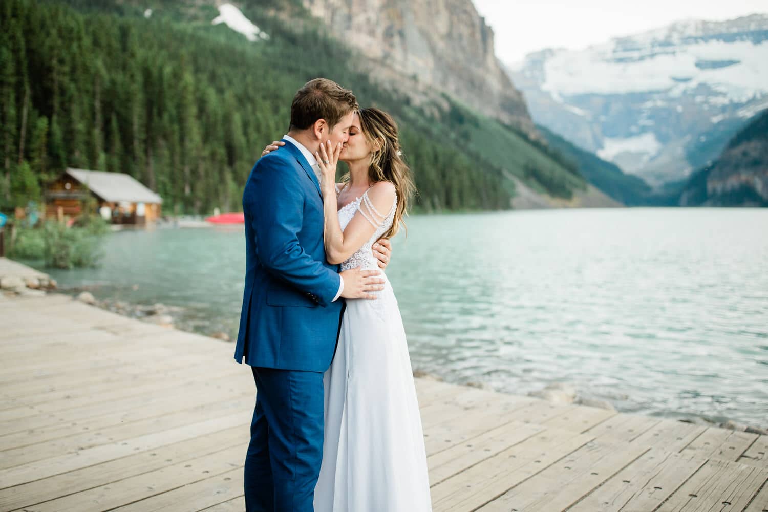 A couple sharing a kiss on a wooden dock by a serene lake, surrounded by mountains and lush greenery.