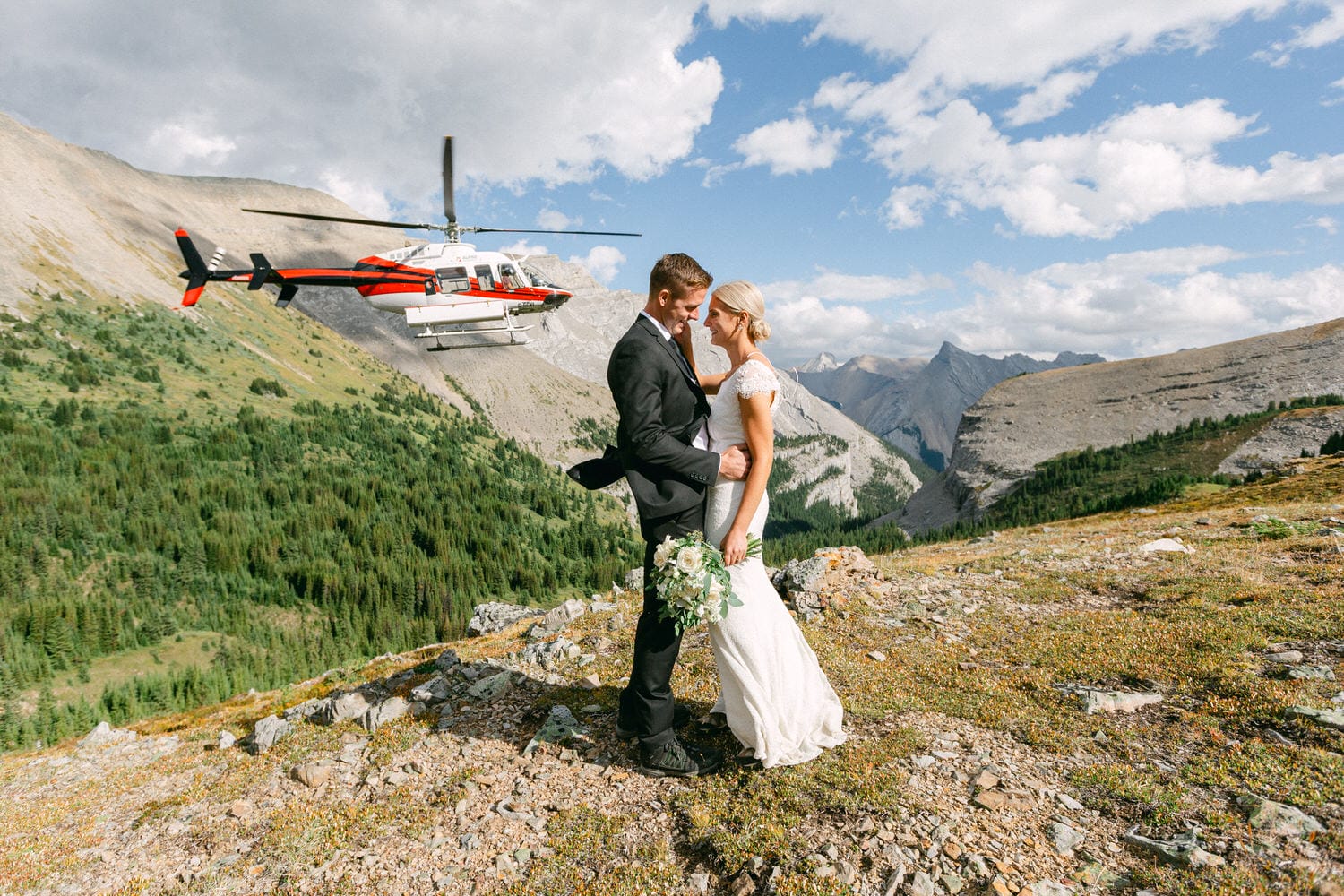 A bride and groom embracing on a rocky mountainside with a helicopter in the background, surrounded by lush greenery and stunning alpine scenery.