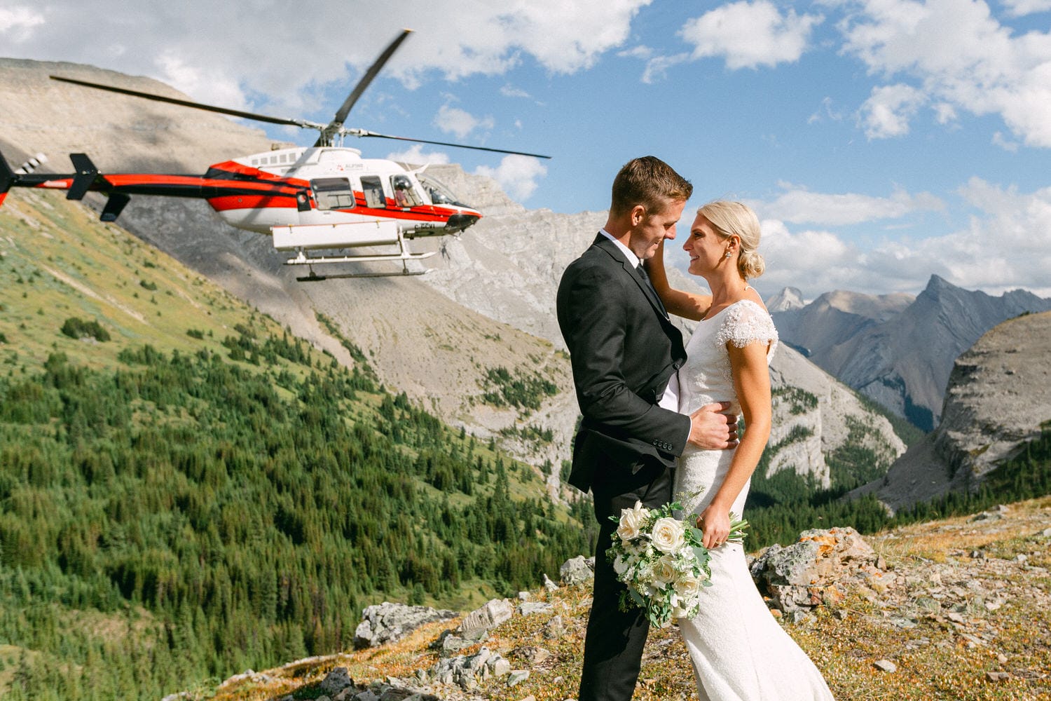 A couple embraces in their wedding attire with a helicopter flying in the background amidst a stunning mountain landscape.