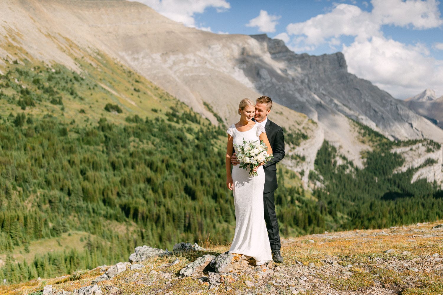 A happy couple in formal attire stands together in a picturesque mountainous landscape, surrounded by lush greenery and rocky terrain, with the bride holding a bouquet.