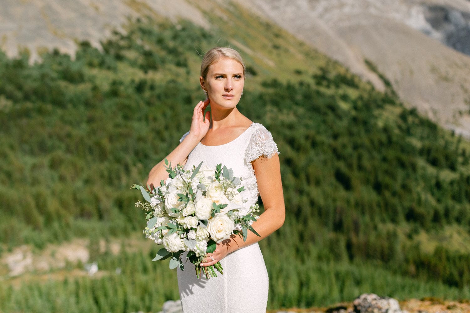 Wedding Bliss in Nature::A bride in a white dress holding a bouquet of white flowers stands against a lush green mountainous backdrop, exuding elegance and serenity.