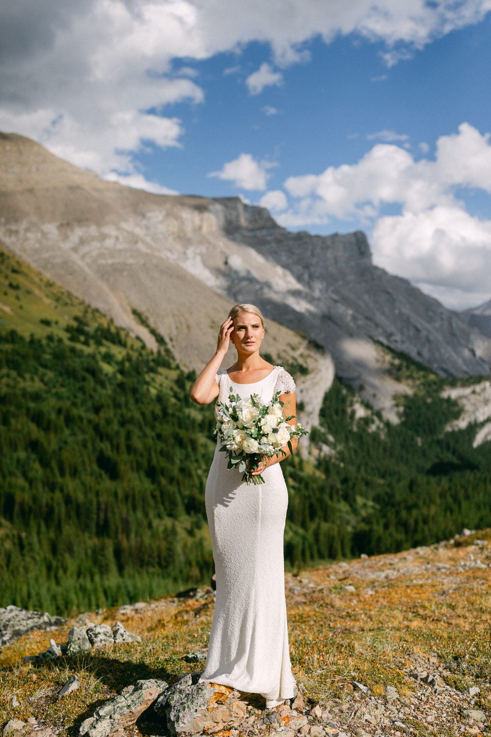 A bride in a white dress stands in a mountainous landscape holding a bouquet of white flowers, with green trees and rocky terrain in the background under a blue sky.