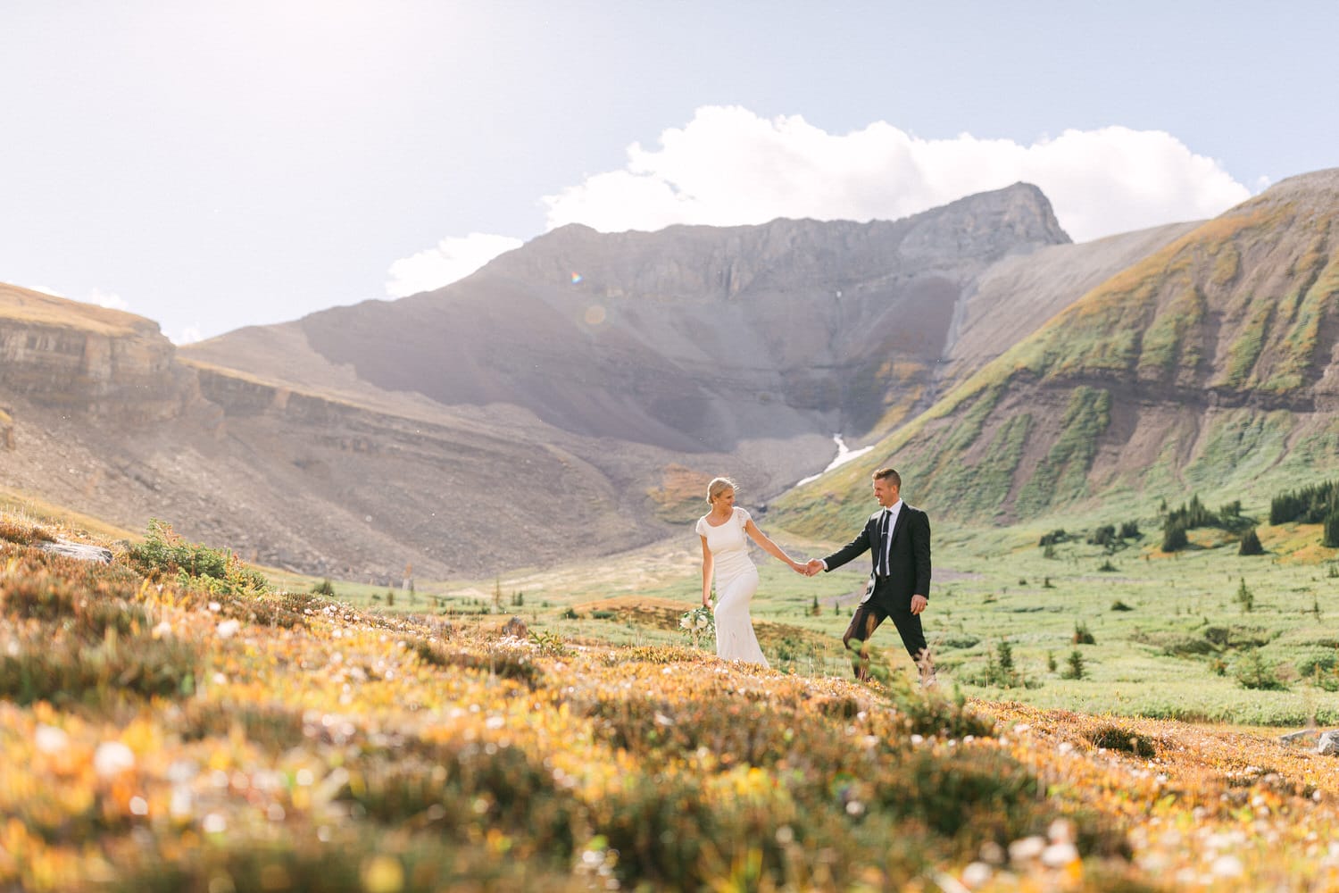 A bride and groom walking hand in hand through a vibrant mountain landscape, surrounded by colorful wildflowers and majestic peaks under a bright blue sky.